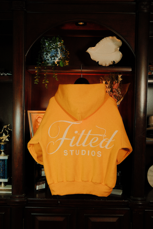 Yellow hoodie with 'Fitted Studios' chain-stitched logo hanging on a wooden shelf.